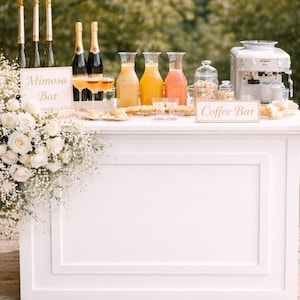 May include: A white mimosa and coffee bar with bottles of champagne, juice carafes, and a coffee machine. Signs read "Mimosa Bar" and "Coffee Bar". The bar is decorated with white roses and baby's breath.