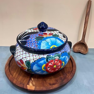 Puede incluir: Un plato de servir grande, redondo, de cerámica azul y blanca con tapa. El plato está decorado con flores coloridas y patrones geométricos. Está sentado en una bandeja de servir de madera.