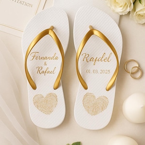 Puede incluir: Chanclas blancas con detalles dorados, personalizadas con nombres y una fecha, y un diseño de huella dactilar en forma de corazón. Las chanclas forman parte de un tema de boda, con una tarjeta de invitación y anillos de boda en el fondo.
