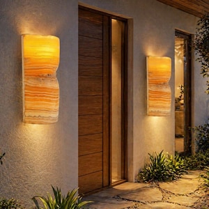Peut inclure: Appliques murales extérieures dégageant une lueur chaleureuse. Les luminaires sont faits d'un matériau ambré en couches et sont montés sur un mur en stuc de couleur claire. Les appliques sont de chaque côté d'une porte en bois.