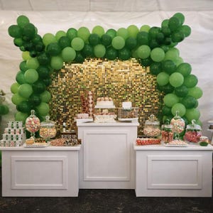 May include: A party setup with white tables, a gold sequin backdrop, and a green balloon arch. The tables display a cake, candies in glass jars, and other treats. People are in the background.