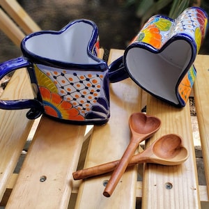 Puede incluir: Dos tazas de cerámica en forma de corazón con esmalte azul y blanco y diseños florales coloridos, junto con dos cucharas de madera en forma de corazón. Las tazas y las cucharas están sobre una superficie de madera.
