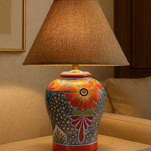 May include: A colourful ceramic table lamp with a floral pattern. The base is painted with orange, blue, and red hues. It has a brown lampshade. The lamp is set on a light-coloured surface.