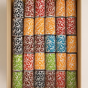 Puede incluir: Una caja rectangular llena de objetos cilíndricos. Cada cilindro está decorado con un patrón floral en varios colores, incluyendo negro, rojo, naranja, amarillo, azul y verde. Los cilindros están dispuestos en filas y columnas ordenadas.