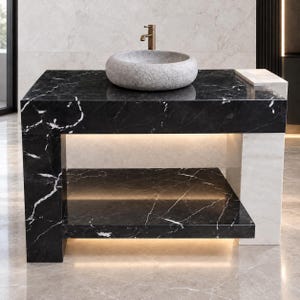 Puede incluir: Un mueble de lavabo de mármol negro con vetas blancas, que incluye un estante inferior e iluminación LED integrada. Un lavabo de piedra gris texturizada se asienta sobre la encimera con un grifo de bronce. Un pilar de mármol blanco sostiene un lado.
