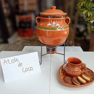 Puede incluir: Una olla de barro con tapa, detalles pintados en verde y blanco, y dos asas, se encuentra sobre un soporte de metal negro. Una taza y un plato a juego con galletas están al lado. El letrero dice "Atole de Coco".
