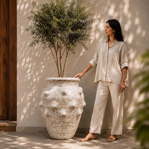 Peut inclure: Un grand pot en céramique blanche texturée avec une finition mouchetée et des protubérances arrondies en relief sur l'extérieur. Le pot contient un grand arbuste aux feuilles vertes et aux branches fines, créant un contraste saisissant.