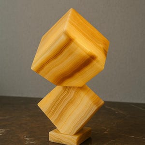 Puede incluir: Una escultura decorativa compuesta por dos formas geométricas apiladas. La escultura está hecha de una piedra de color claro con vetas marrones cálidas. La base es cuadrada y la parte superior es un cubo, creando un diseño abstracto moderno.