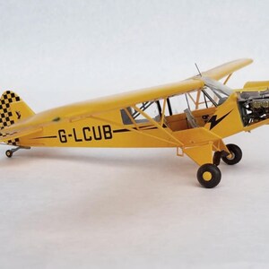 Puede incluir: Un modelo de avi&oacute;n amarillo con texto negro "G-LCUB" en el lateral. La cola presenta un patr&oacute;n de cuadros blancos y negros. El avi&oacute;n tiene una h&eacute;lice y ruedas negras. El modelo est&aacute; sobre un fondo blanco.