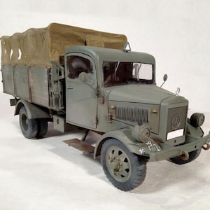 Może przedstawiać: Szczegółowy model zabytkowej ciężarówki wojskowej w kolorze oliwkowym. Ciężarówka posiada plandekę na skrzyni ładunkowej, klasyczny wygląd kabiny oraz widoczny emblemat Mercedes-Benz z przodu. Model umieszczono na gładkim tle.