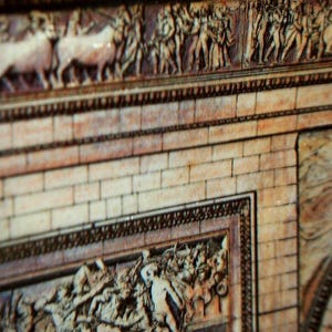 May include: Close-up of a section of a detailed relief sculpture, featuring figures and animals. The relief is carved into a light-colored material, with a brick-like pattern below. The artwork has a classical style, with intricate details and a sense of depth.