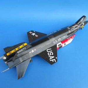 May include: A black and gray model airplane with the text "NASA" and "U.S. Air Force". The model has red and white accents and the number "66671". The wings have white stars.