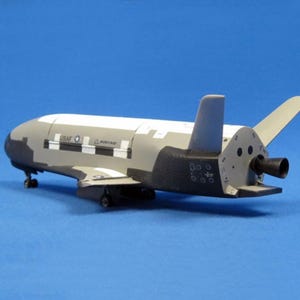以下が含まれることがあります： グレーと白を基調とした宇宙船の模型で、黒いアクセント、翼、尾翼があります。モデルには「USAF」と「Boeing」の文字が印刷されています。宇宙船は青色の背景に配置されています。