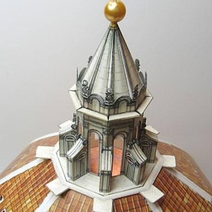 May include: A paper model of a dome with a gold-coloured orb and cross on top. The dome is made of white paper with black lines and details. The base of the dome is made of brown paper with a chequered pattern.