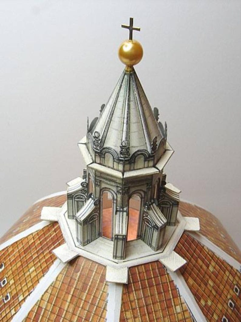 May include: A paper model of a dome with a gold-coloured orb and cross on top. The dome is made of white paper with black lines and details. The base of the dome is made of brown paper with a chequered pattern.