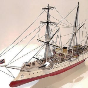 Puede incluir: Un modelo detallado de un barco de vela blanco y rojo con aparejos negros y una bandera de la Unión Jack. El barco tiene múltiples mástiles y velas, y un casco rojo con detalles blancos.