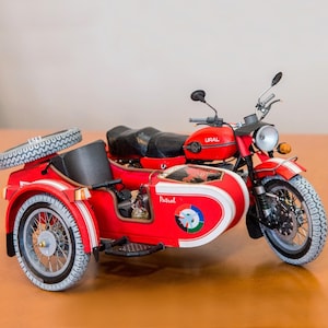 Può includere: Una moto rossa con sidecar. La moto ha una sella nera, una ruota di scorta e la scritta "URAL" sul serbatoio. Il sidecar è rosso con dettagli bianchi e la scritta "Petrol". I pneumatici sono neri e bianchi.