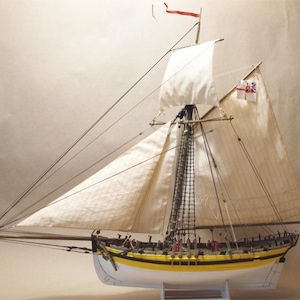 May include: A detailed model sailboat with white sails, a yellow and black hull, and rigging. The model features a British flag and a red pennant. The boat is displayed on a white stand, showcasing its intricate design and craftsmanship.