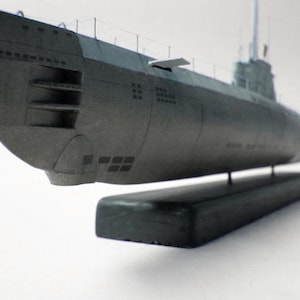 Puede incluir: Un modelo de submarino gris sobre una base oscura. El submarino tiene una proa redondeada, una torre de mando y varias ventanas rectangulares. El modelo se exhibe sobre una base rectangular negra.