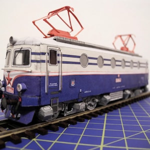 Op de afbeelding: Een gedetailleerd model van een treinlocomotief in blauw, wit en rood. Het model heeft een rood sterembleem en staat op een miniatuurbaan. De trein heeft een gedetailleerd ontwerp met zichtbare wielen en bovenleidingapparatuur.