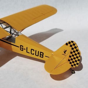 Puede incluir: Un modelo de avi&oacute;n amarillo con cola a cuadros blancos y negros y el texto "G-LCUB". El avi&oacute;n tiene una envergadura de aproximadamente 25 cm. El modelo es una r&eacute;plica de un avi&oacute;n antiguo.