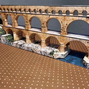 Könnte beinhalten: Ein detailliertes Modell einer antiken römischen Aquäduktbrücke mit mehreren Bögen und Ebenen. Die Struktur besteht aus beigefarbenem Stein, mit einem blauen Fluss, der darunter fließt. Das Modell steht auf einer braunen Oberfläche mit weißen Punkten.