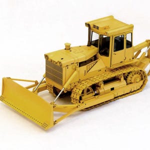 Op de afbeelding: Een geel bulldozer model met een blad, rupsbanden en een cabine. Het bouwmachine staat tegen een witte achtergrond. Het model heeft een gedetailleerd ontwerp, inclusief de radiatorgrille en uitlaat.