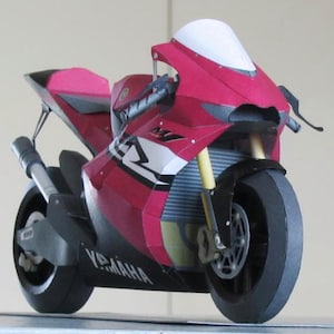 以下が含まれることがあります： ヤマハのオートバイの詳細なペーパークラフトモデル。このスポーツバイクは、赤と黒の配色に白いアクセント、黒いタイヤ、ヤマハのロゴが特徴です。このモデルには、フロントガラスと詳細なエンジンが付いています。