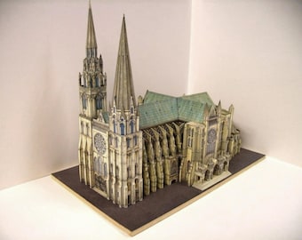 May include: A detailed paper model of a cathedral with two tall spires and intricate details. The model is made of paper and is painted in shades of gray, white, and brown.