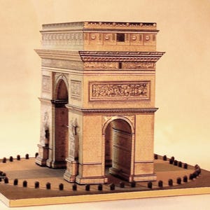 May include: A miniature replica of the Arc de Triomphe, a famous Parisian monument. The model is constructed from tan-colored materials, featuring detailed architectural elements, arches, and sculpted reliefs. The base includes a circular area with small, dark posts.