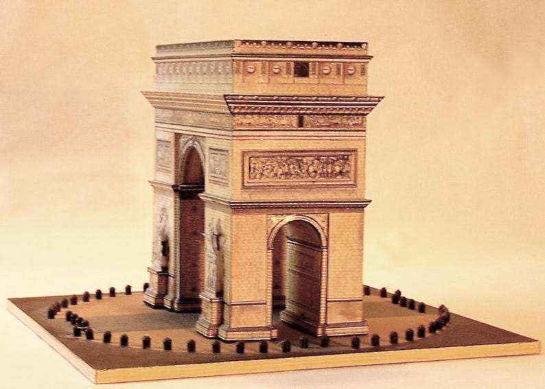 May include: A miniature replica of the Arc de Triomphe, a famous Parisian monument. The model is constructed from tan-colored materials, featuring detailed architectural elements, arches, and sculpted reliefs. The base includes a circular area with small, dark posts.