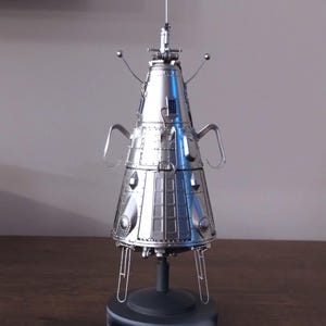 May include: A detailed, silver-colored model of a spacecraft, possibly a capsule or satellite. The model has a conical shape with multiple panels, antennas, and support legs. It is displayed on a dark gray base, showcasing intricate details.