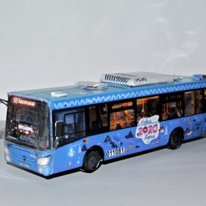 Könnte beinhalten: Ein blaues Spielzeugbusmodell mit einem weißen Zickzackmuster entlang der Dachlinie. Der Bus trägt den Text "2020" und andere Texte in einer Fremdsprache. Das Modell hat die Nummer "011081" an der Seite.