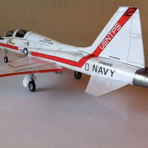 May include: A white and red model aeroplane with the text "USNTPS" and "NAVY" on the tail. The model is on a white surface.