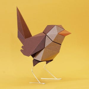 May include: A geometric paper bird sculpture in shades of brown and tan, with an orange beak and wire legs, set against a yellow background. The bird's form is constructed from folded paper, showcasing a modern, minimalist design.