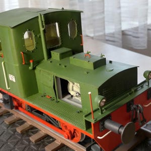 以下が含まれることがあります： 緑と赤の色合いの、詳細なモデルの列車機関車が線路の一部に置かれています。モデルは窓付きのキャブ、詳細なエンジンコンパートメント、目立つフロントバンパーを備えています。モデルの長さは約30cmです。