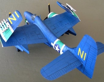 May include: A blue and green model airplane with the letters "NI" on the tail and wings. The plane has a star on the side and is sitting on a light brown surface.