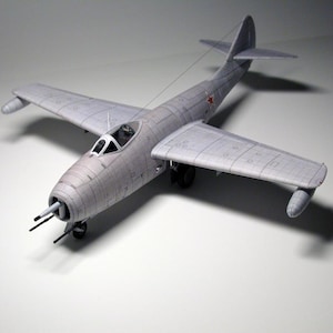 May include: A gray paper model of a Soviet MiG-15 jet fighter aircraft. The model is detailed with a cockpit, wings, and tail.