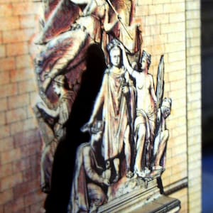 May include: A detailed relief sculpture depicting classical figures, set against a brick wall background. The artwork features multiple figures in draped garments, with a central figure holding a staff. The color palette includes beige, brown, and hints of red.