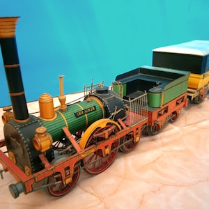 Könnte beinhalten: Ein detailliertes Modell eines Zuges in Schwarz, Grün und Orange. Die Lokomotive hat einen hohen Schornstein und die Aufschrift "DER ADLER". Der Zug umfasst einen Personenwagen mit blauem und gelbem Dach.