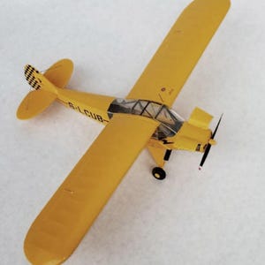Puede incluir: Un modelo de avión amarillo con hélice y ruedas negras. El avión tiene una cabina transparente y el texto "G-LCUB" en el lateral. La cola presenta un patrón de cuadros blancos y negros.
