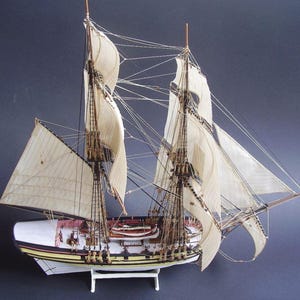May include: A detailed model of a sailing ship with white sails and intricate rigging. The ship's hull is painted white, with yellow and maroon accents. The model is displayed on a white stand against a dark blue background.