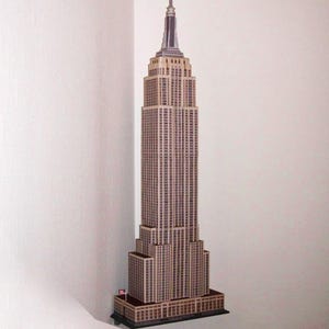 Può includere: Un modello dettagliato dell'Empire State Building, nei toni del beige e del marrone. Il modello ha un design a gradini con un'alta guglia e una piccola bandiera americana. È posto in un angolo contro un muro bianco.