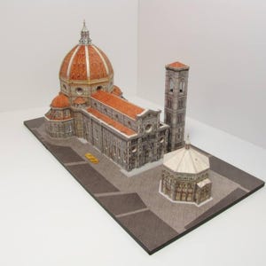 May include: A detailed model of the Florence Duomo, a famous Italian cathedral, with a red tiled roof, a large dome, and a bell tower. The model is made of paper and is displayed on a brown base.