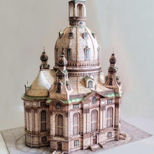 May include: A detailed paper model of a church with a large dome and multiple spires. The model features intricate architectural details, including arched windows, columns, and a textured facade in shades of beige, brown, and green.