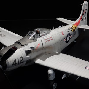 May include: A detailed model airplane in shades of gray and white, featuring a propeller, wings, and tail. The model has the number "412" on the fuselage and "AK 35326" on the tail. A stylized bee graphic is visible on the tail.