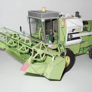 May include: A green and white paper model of a combine harvester. The model has a detailed design with a large header and a small cab. The model is made of paper and is a great example of a papercraft project.
