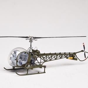 May include: A detailed model helicopter with a clear bubble cockpit and olive drab frame. The model features a main rotor, tail rotor with red tips, and landing skids. The overall design is a miniature representation of a military helicopter.