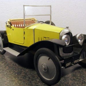 以下が含まれることがあります： 黒いフェンダーと車輪が付いた、ヴィンテージの黄色の玩具車。車内は茶色で、フロントガラスのフレームはシルバーです。「5HF-1925」と書かれたナンバープレートが付いています。