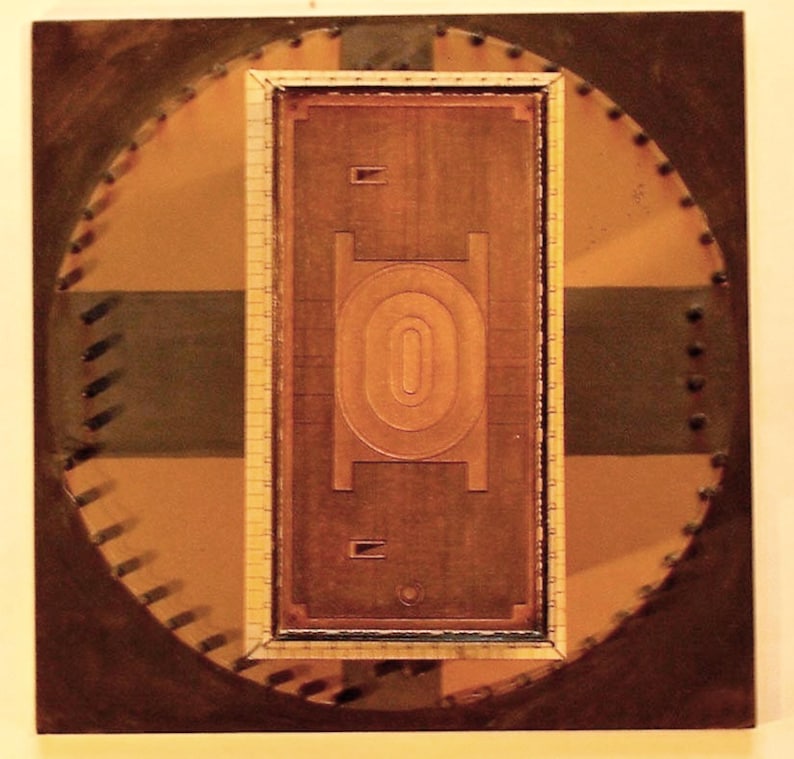 May include: Abstract artwork featuring a rectangular, copper-toned central design with an oval shape, framed within a circular pattern. The artwork incorporates shades of brown, tan, and green, with a textured appearance and rivet-like details around the edges.